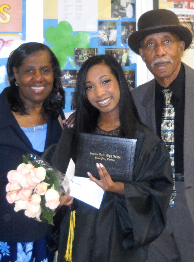Smiling graduate in a cap and gown holds her diploma, flanked by two adults (woman with pink roses, man in a hat) in front of a bulletin-board backdrop.