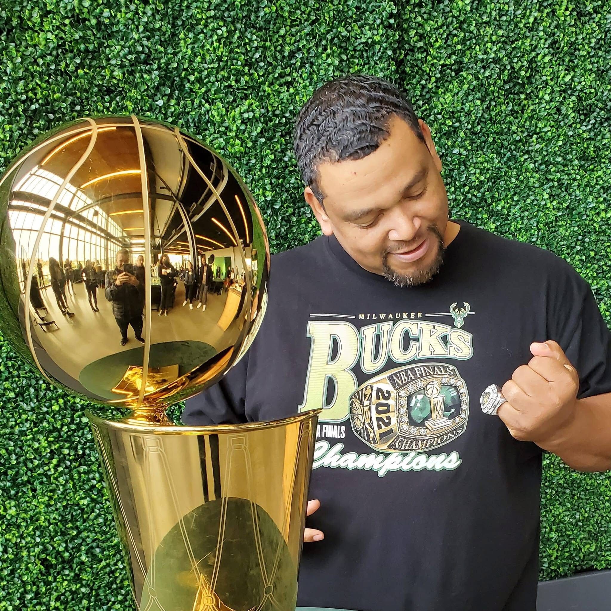 Man posing with a large gold trophy in front of a green hedge wall.