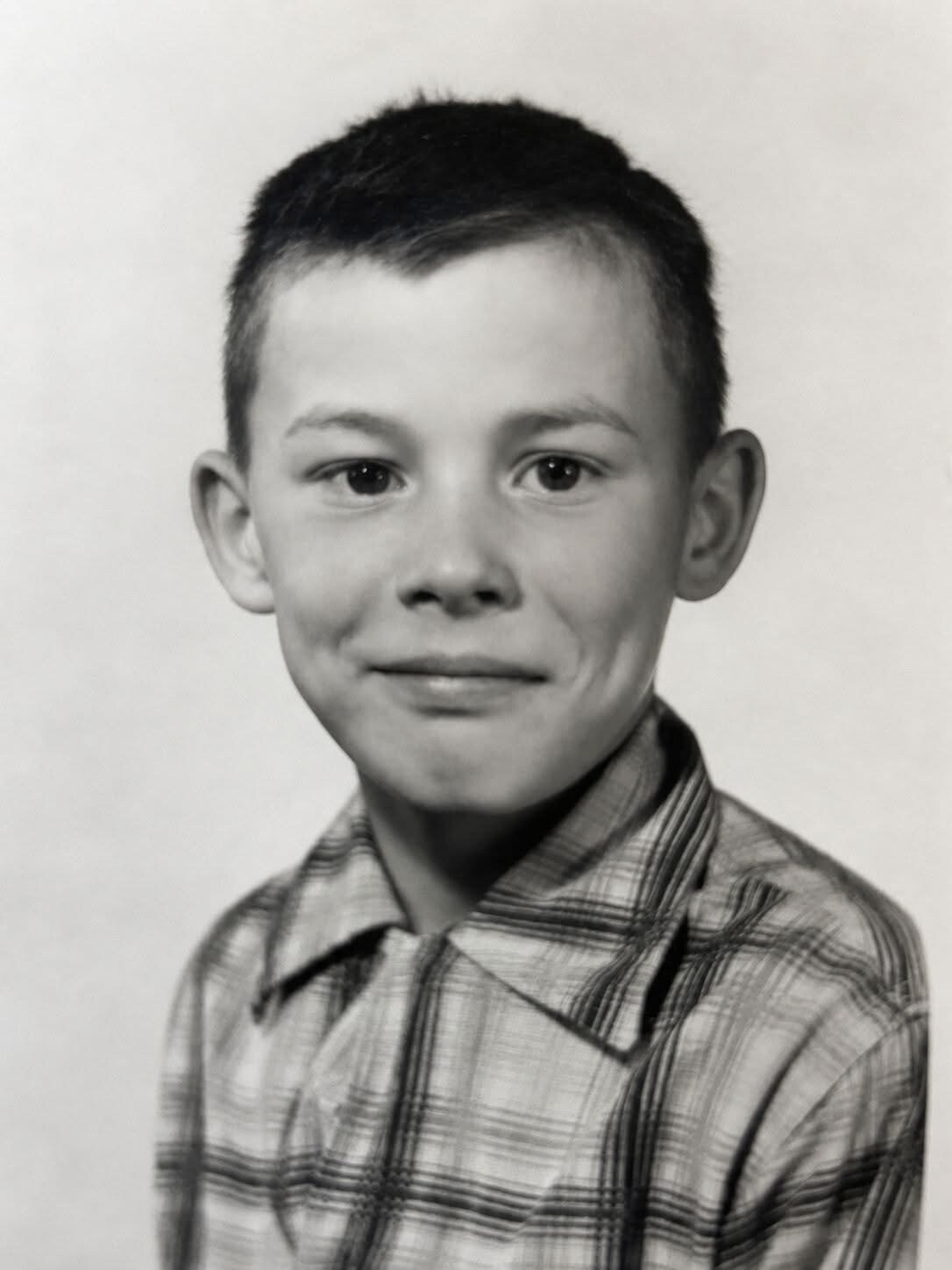 Black-and-white portrait of a young boy with short dark hair, wearing a plaid shirt and a subtle smile, looking at the camera by a neutral backdrop.