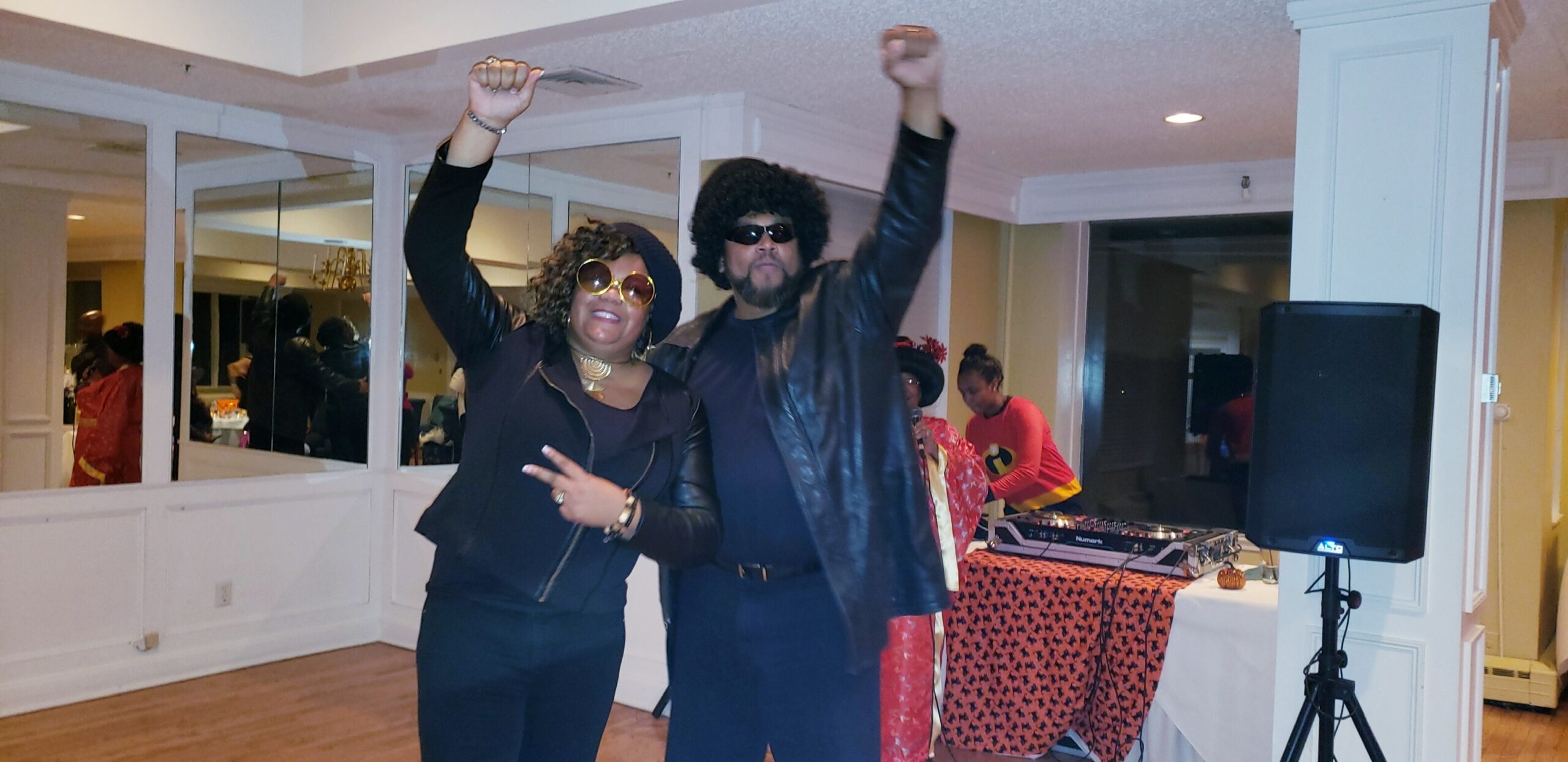 Two people pose and dance at a retro-themed party with a DJ in the background, arms raised in celebration.