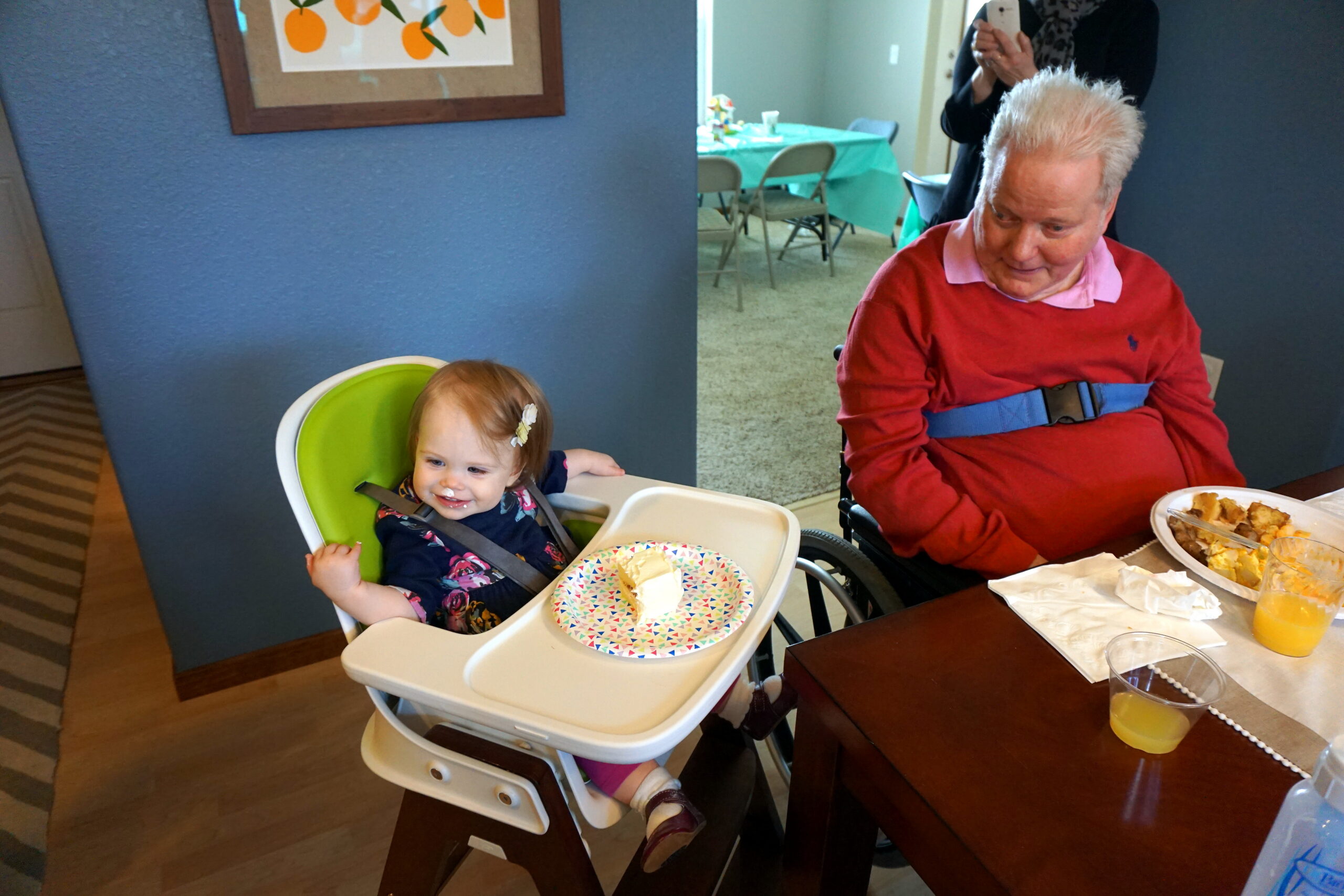 Smiling toddler in a high chair sits beside an elderly man in a wheelchair at a dining table with plates of cake and orange juice nearby.