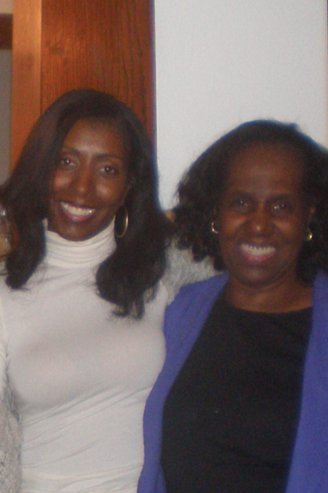 Two women smiling at the camera indoors, one in a white turtleneck and the other in a purple blazer with a black top beneath.