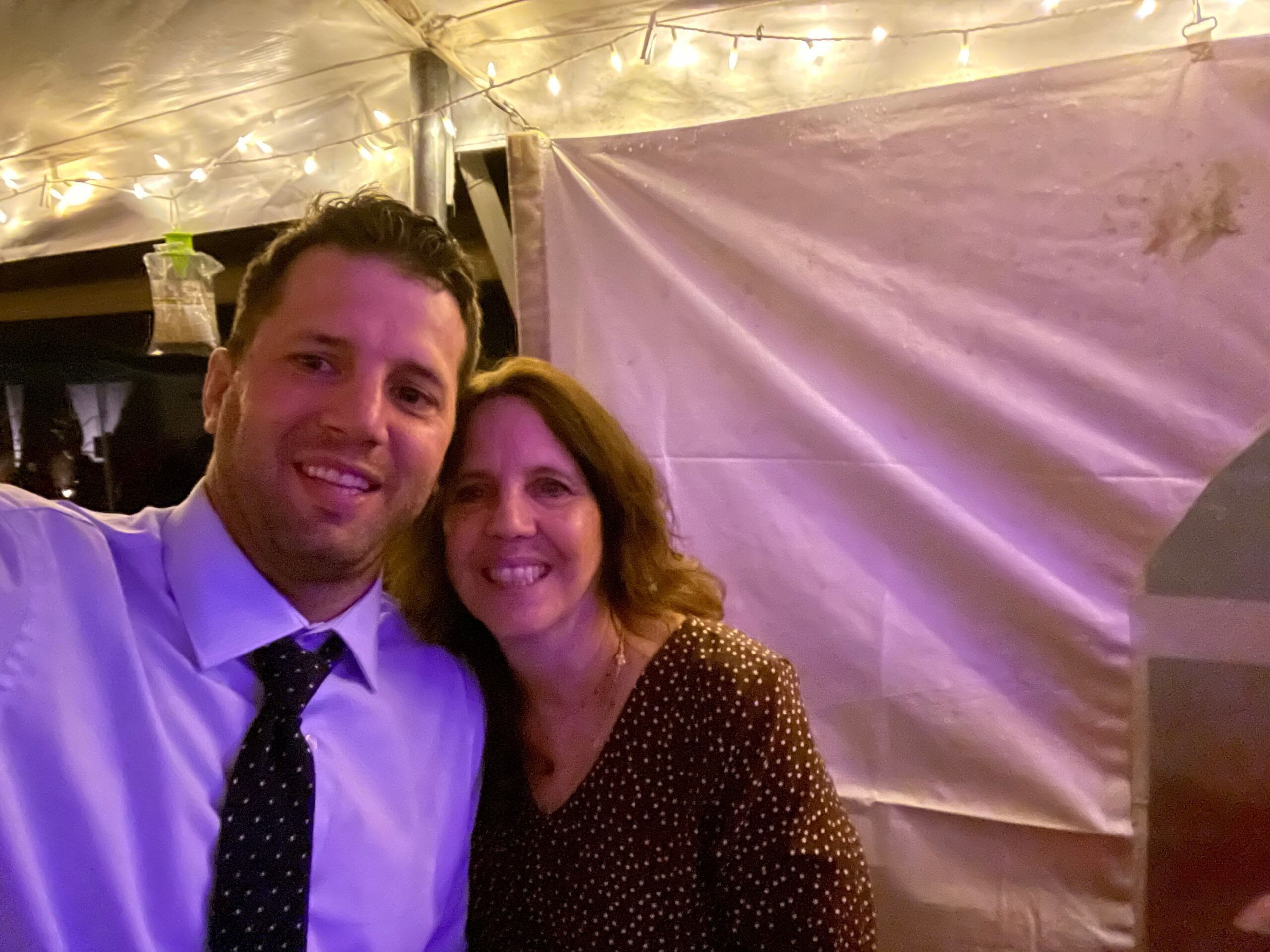 Smiling man in a white shirt and black polka-dot tie takes a selfie with a woman in a brown polka-dot dress under warm string lights in a tent.