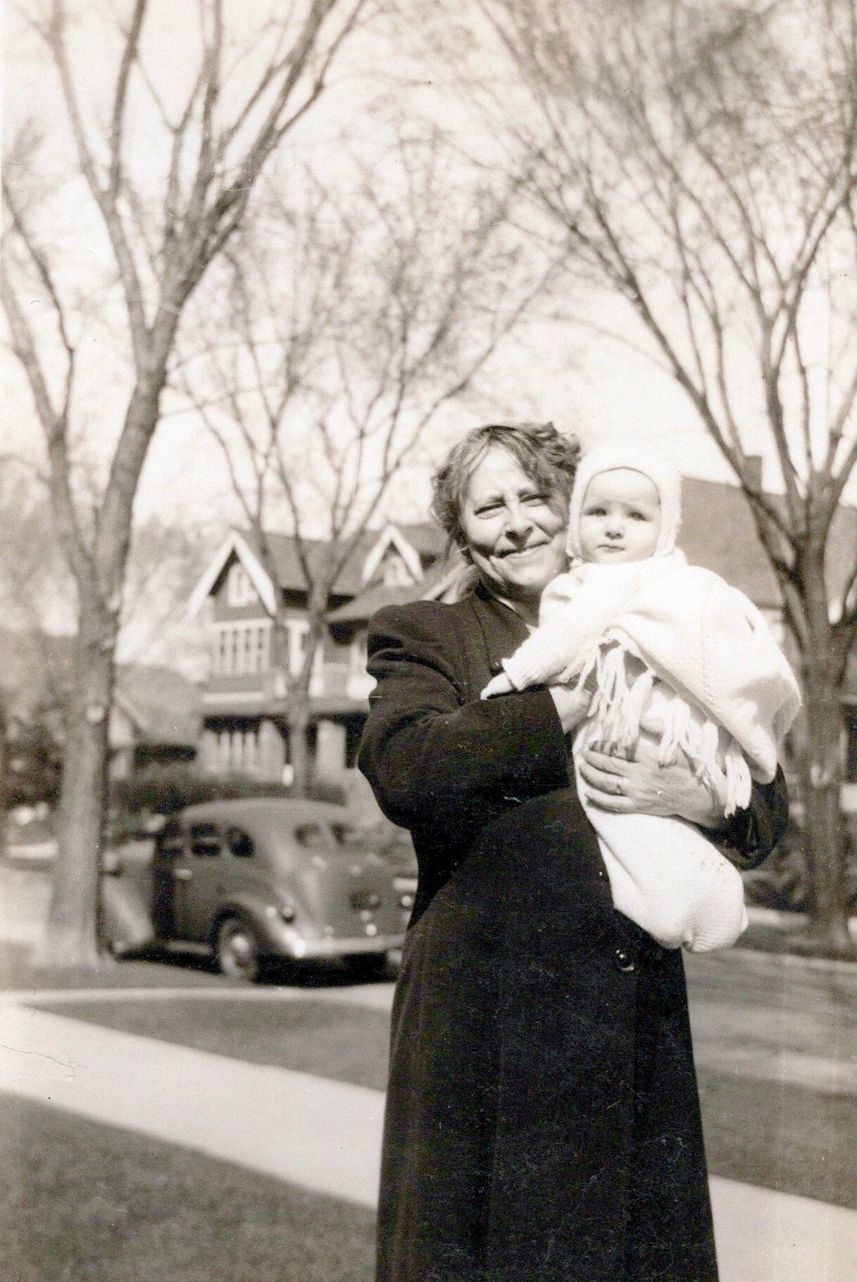 Older woman in a dark coat holding a bundled baby outside, with bare trees and a vintage car in the background.
