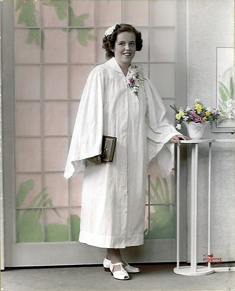 Woman in a white graduation gown holds a book, posing beside a small pedestal with flowers.
