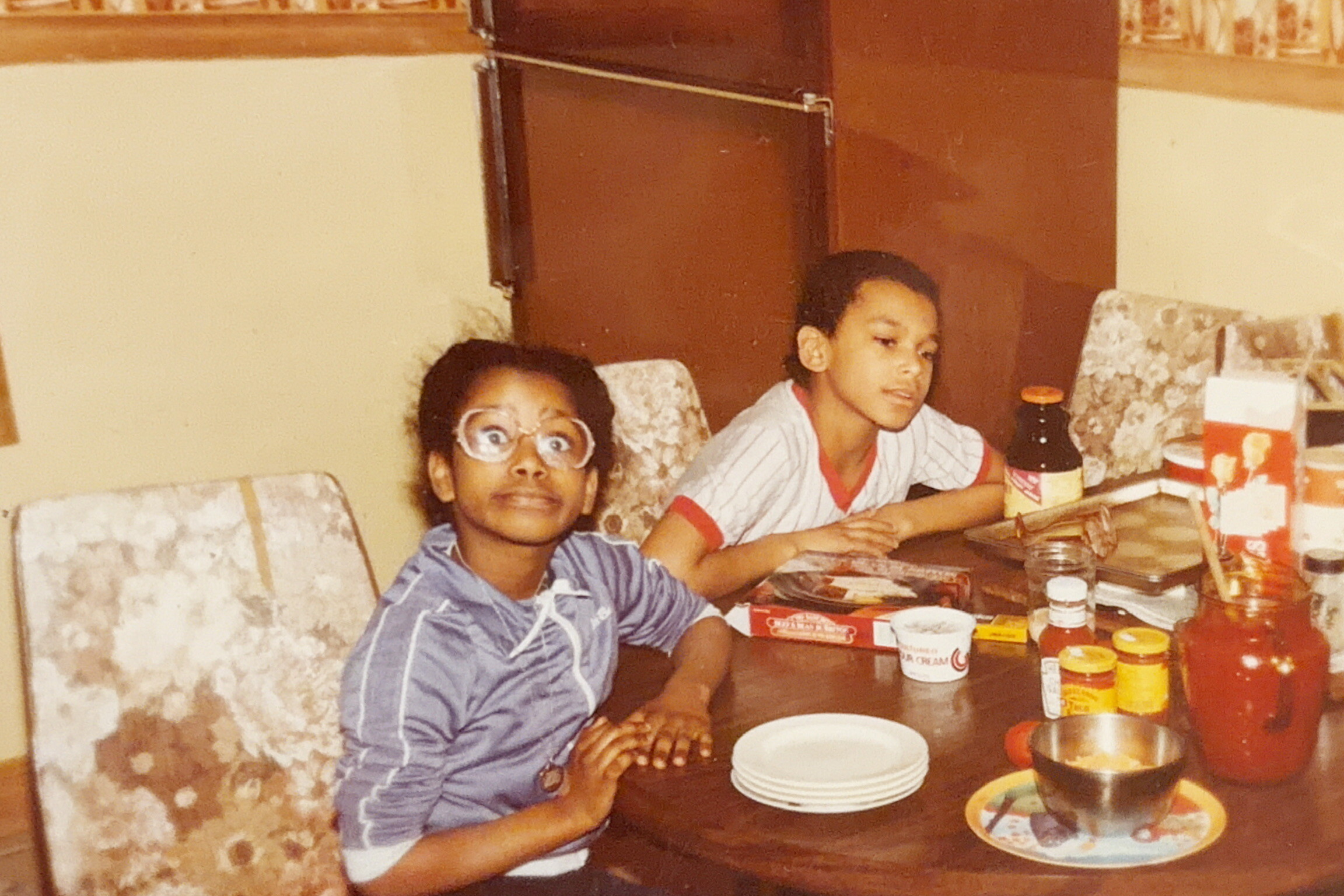 Two boys sit at a dining table with food and condiments, one wearing glasses and a surprised expression as they pose for the photo.
