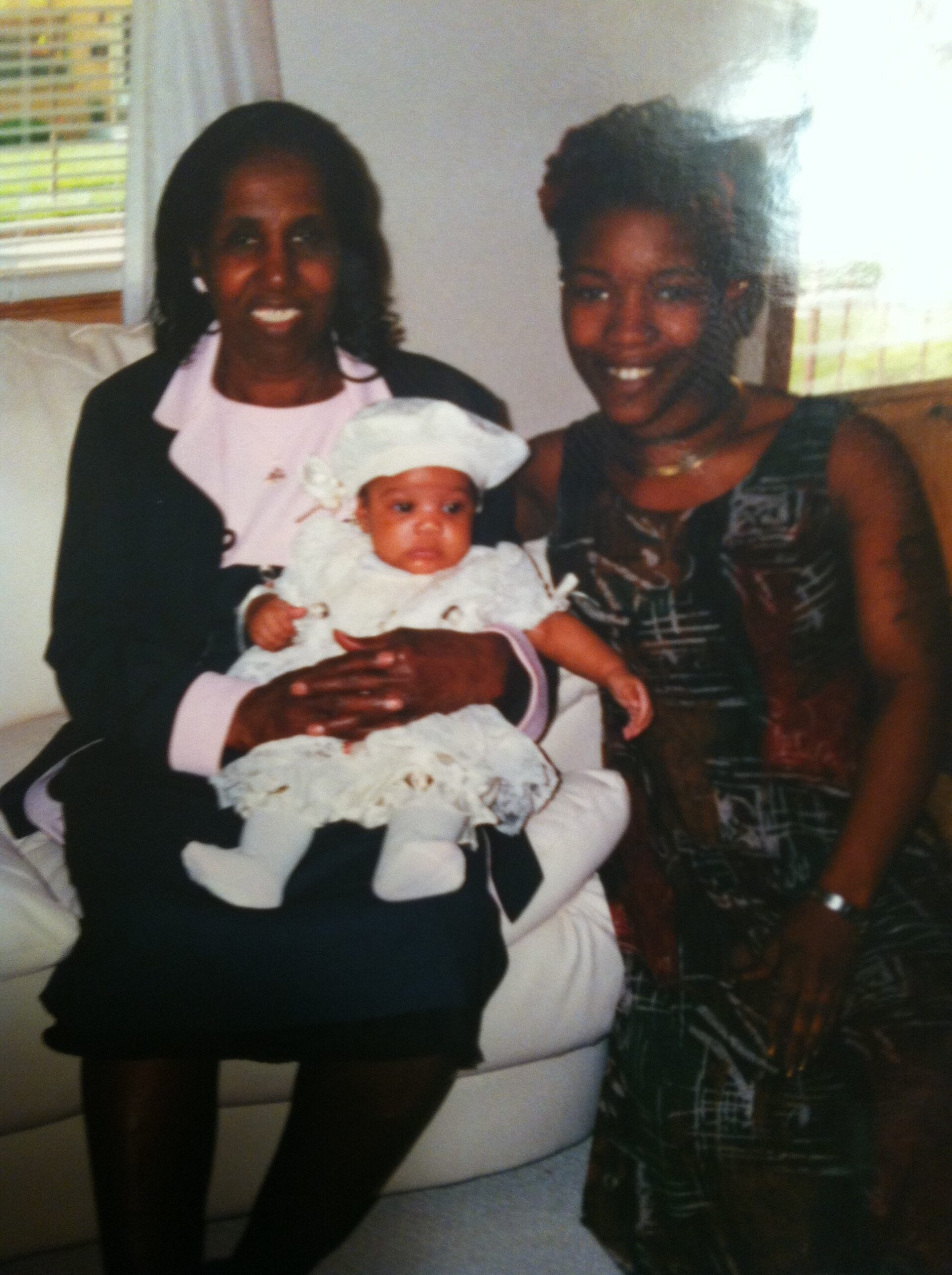 Two women smiling with a baby in white on a sofa in a living room.