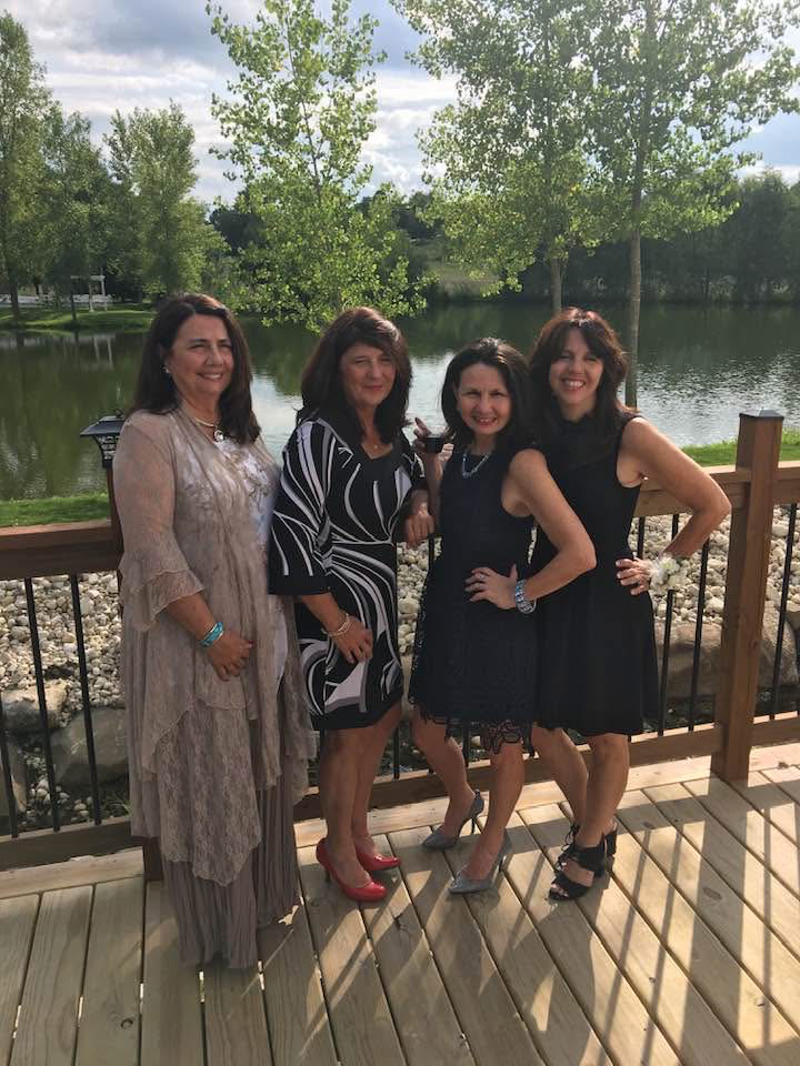 Four women standing on a wooden deck by a lake, posing and smiling with trees in the background.