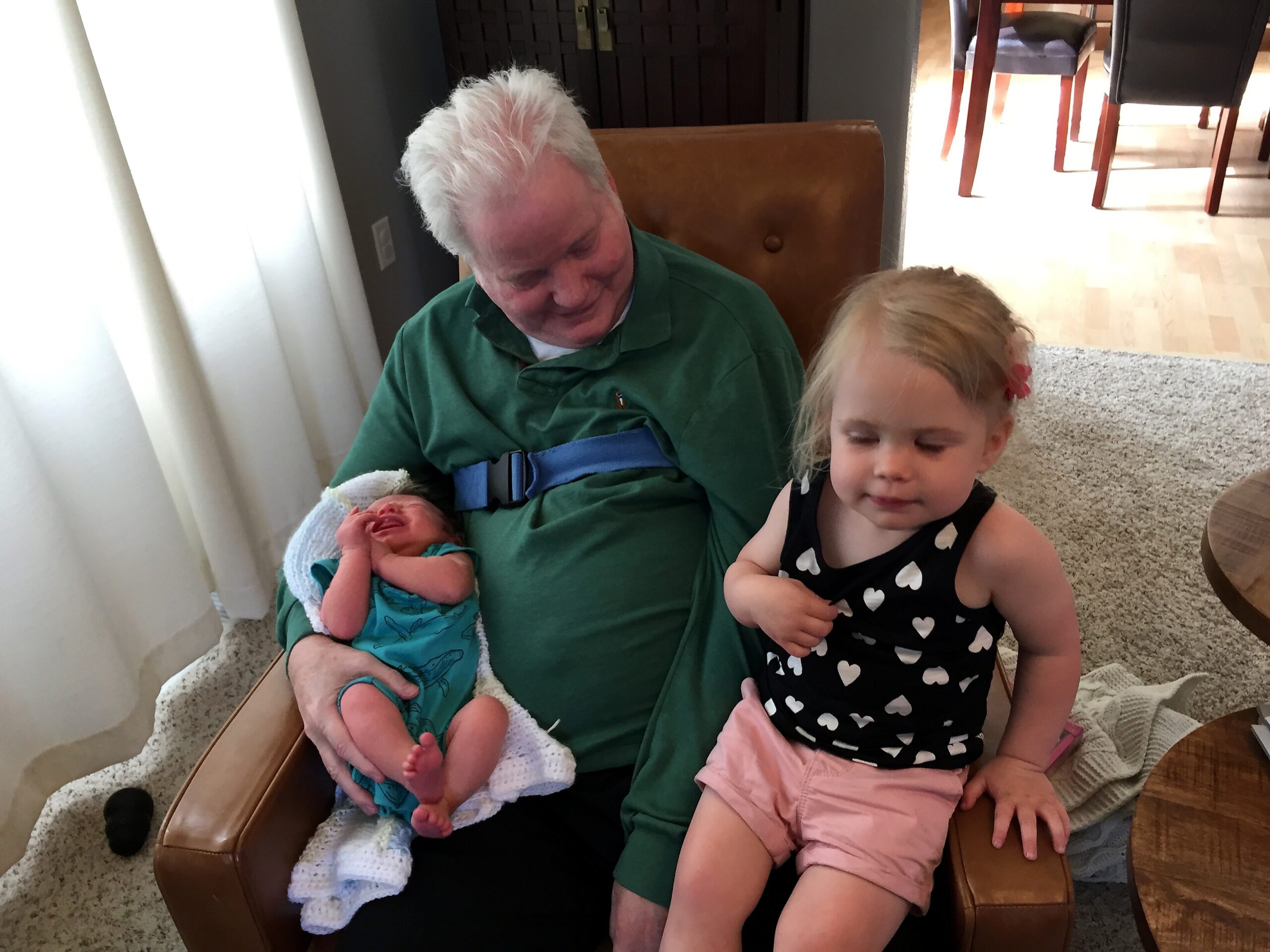 Older man in a green shirt cradles a newborn baby wrapped in a blue blanket, with a young girl in a heart‑print top nearby in a living room.