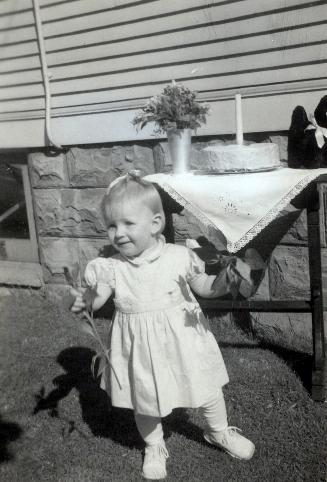Toddler girl in a light dress standing on the lawn, holding a flower stem with a cake and vase of flowers on a table behind her.