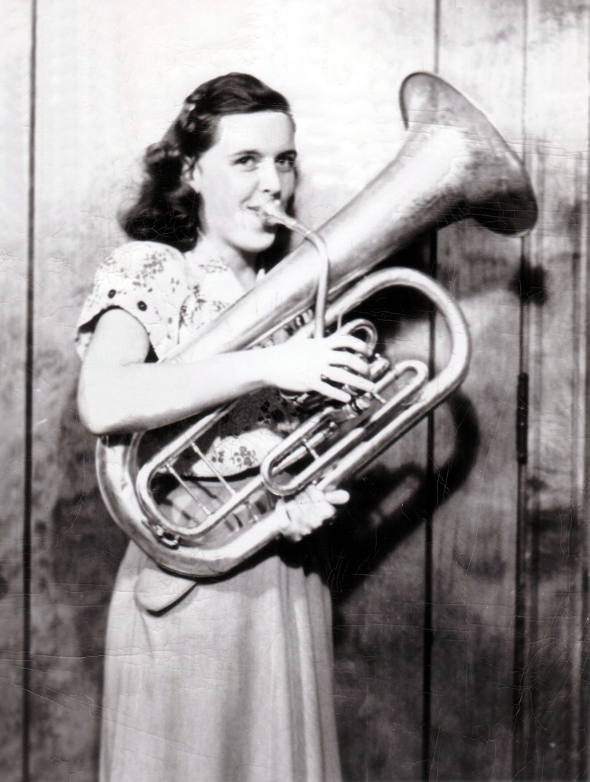 Young girl in a light patterned dress playing a French horn and smiling at the camera in a wooden-room setting.