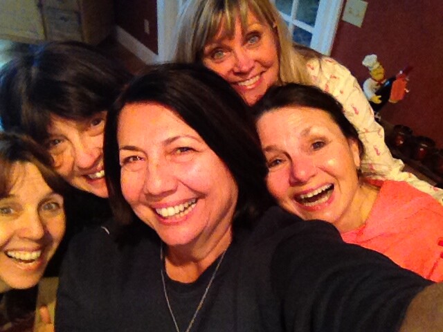 Group of five women taking a close-up selfie, all smiling at the camera indoors at home.