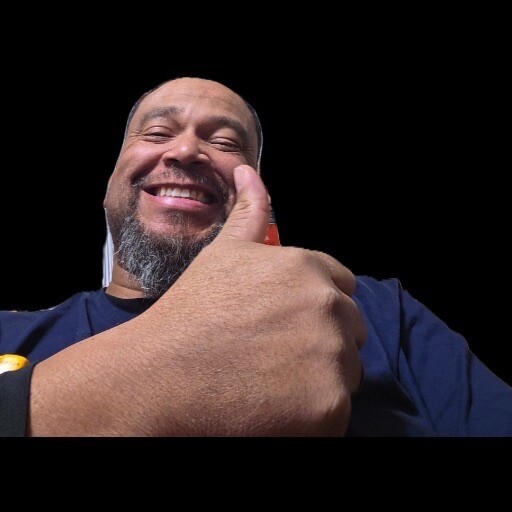 Smiling man giving a thumbs-up toward the camera against a black background.