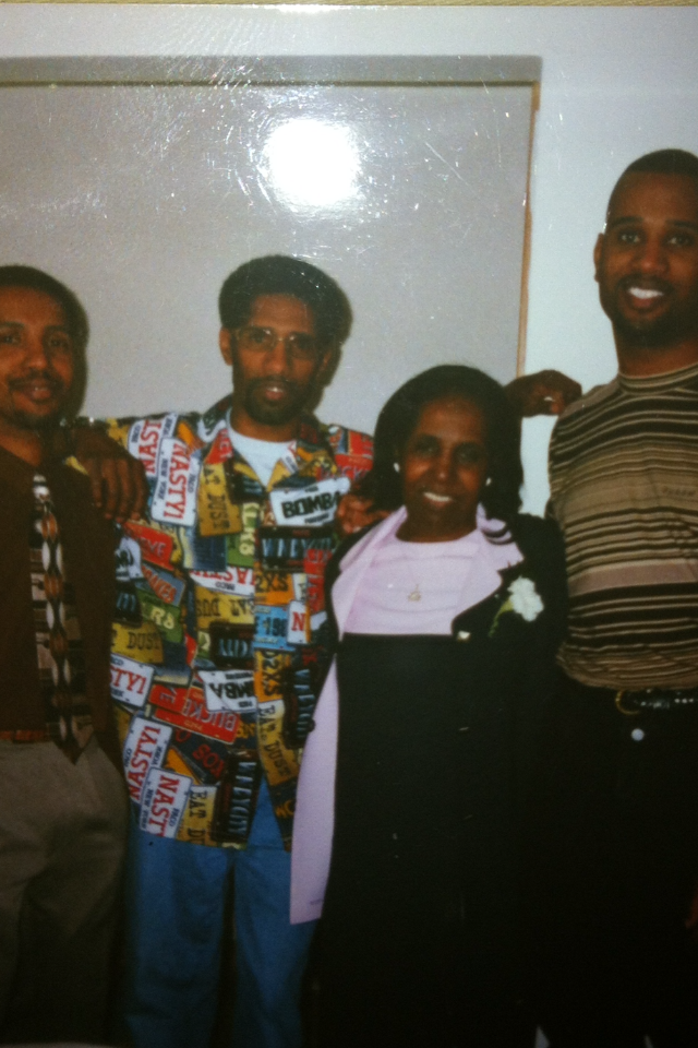 Four adults posing together indoors, smiling at the camera; the man second from the left wears a colorful patchwork jacket covered in logos.