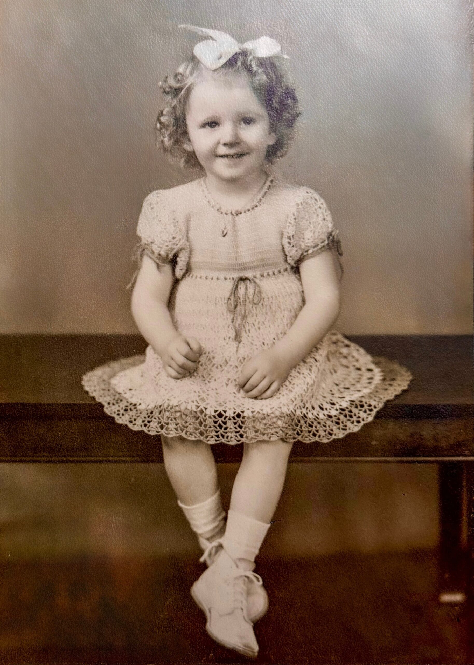 Vintage photo of a young girl with a large bow in her curly hair, wearing a crocheted dress and hat, smiling at the camera.