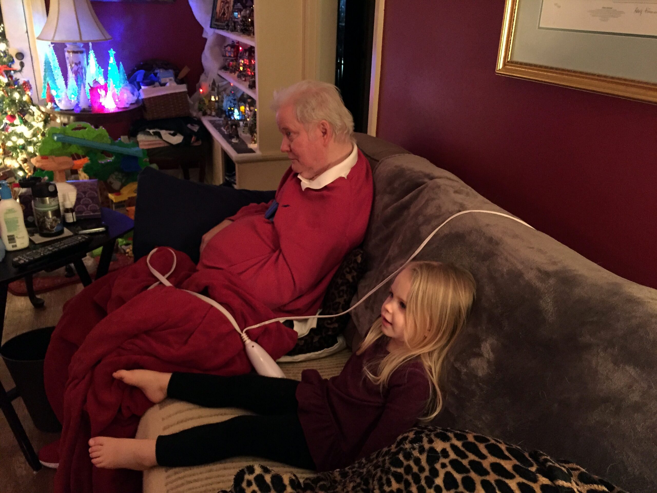 Elderly man in a red robe sitting on a couch under a red blanket, with a young girl resting beside him in a cozy, Christmas-decorated living room.