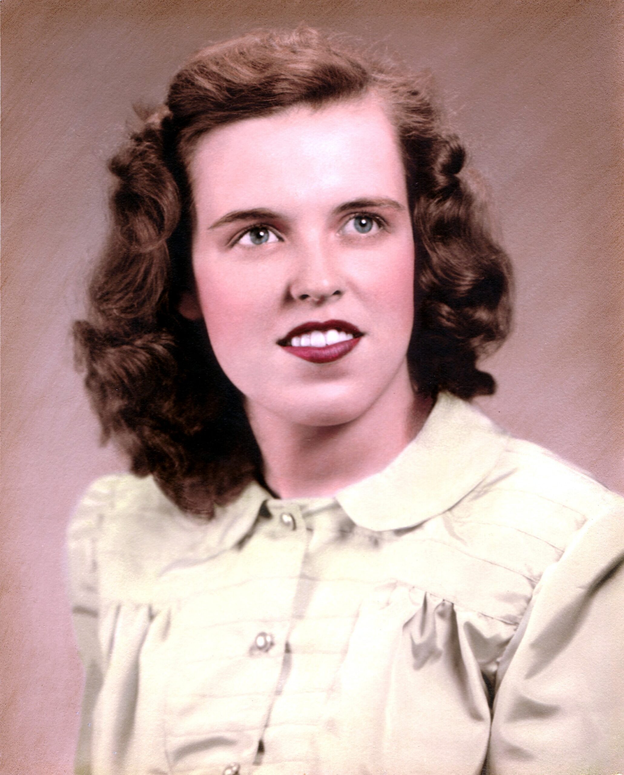 Portrait of a young woman with wavy brown hair and blue eyes, wearing a light collared blouse against a pink studio backdrop.