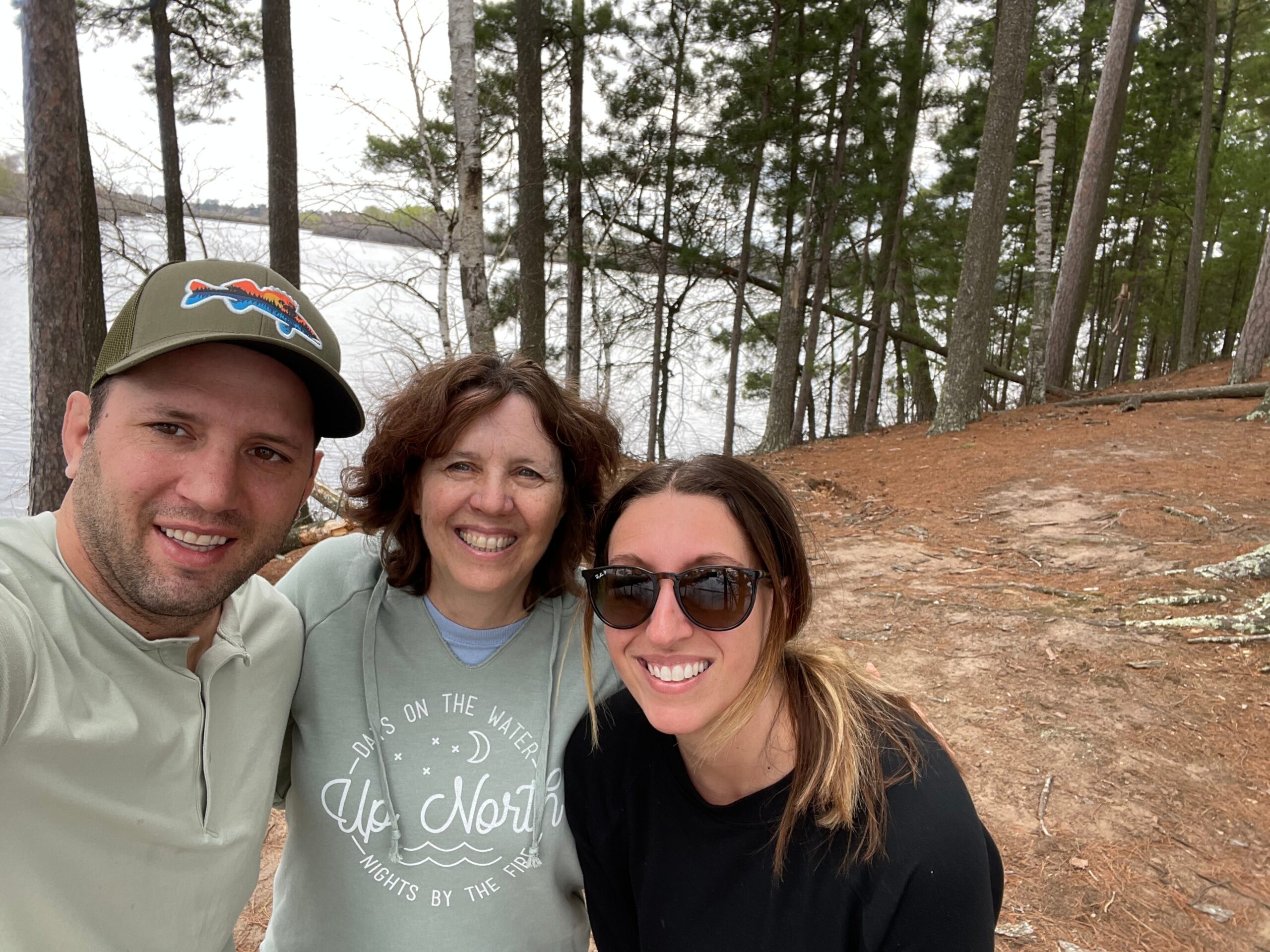 Smiling group of people taking a selfie in a wooded lakeside area with tall trees and a calm lake in the background.