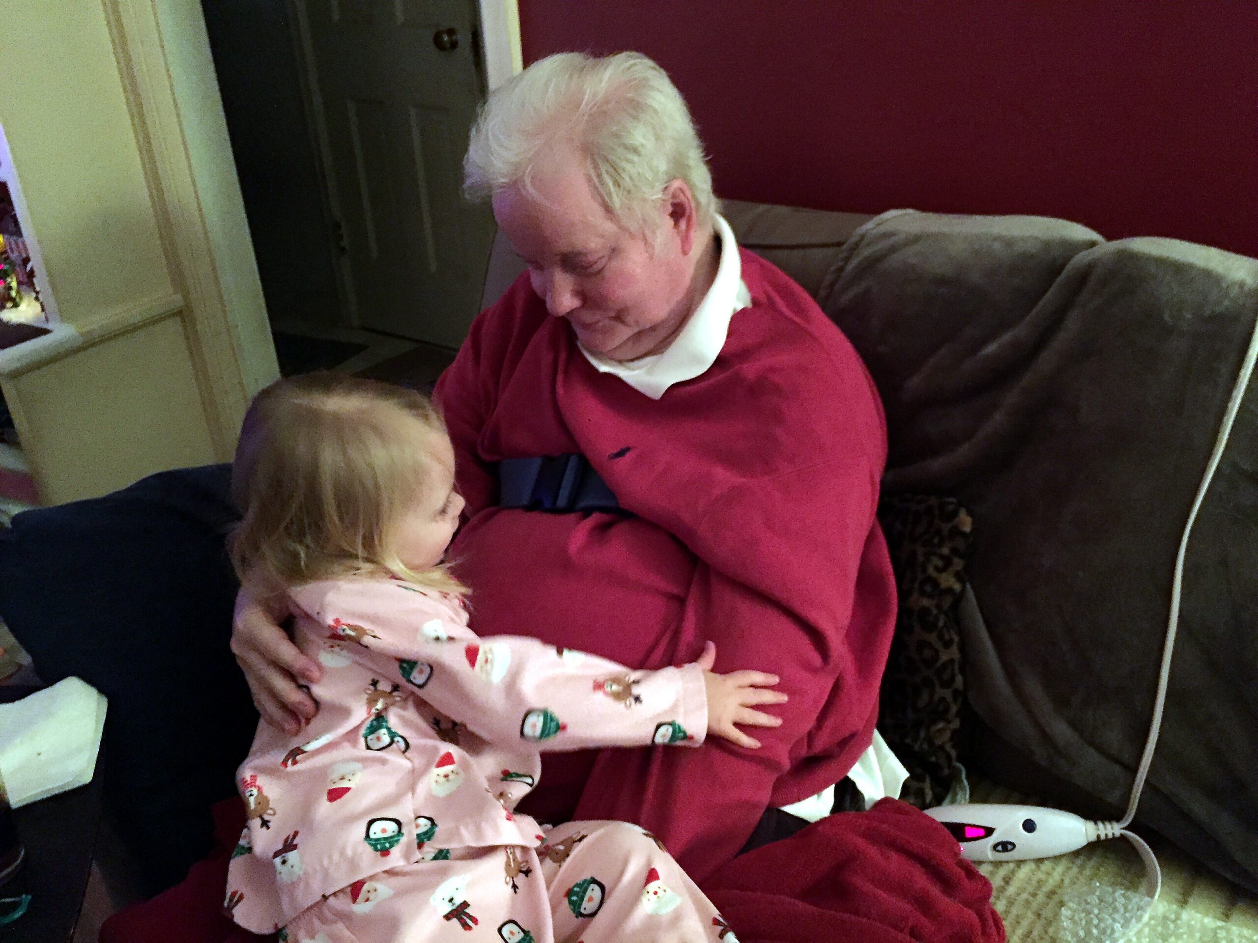 Older man with white hair in a red sweater sits on a couch, holding a young child in pink pajamas.