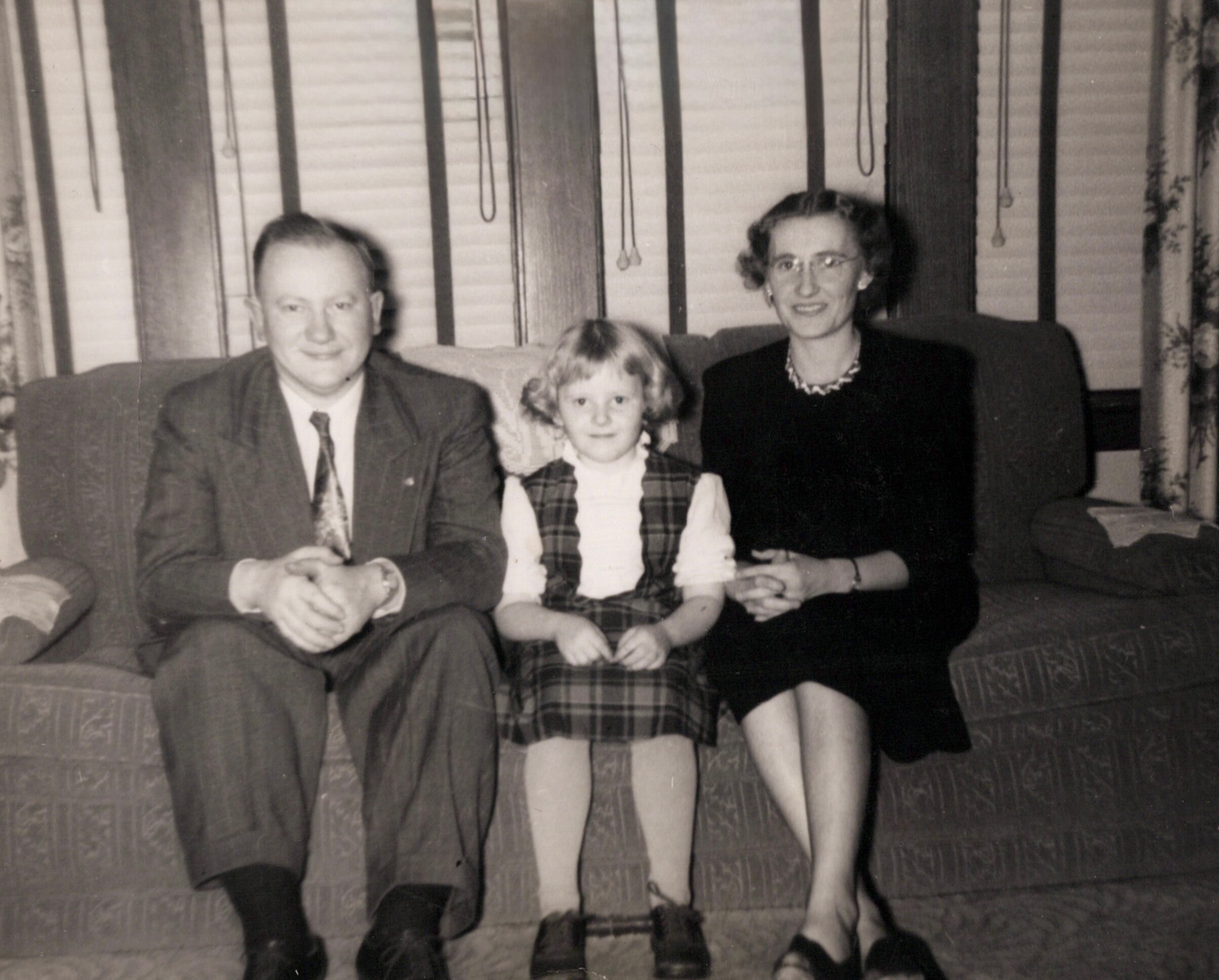 Three generations posing on a sofa: man on left, young girl in middle, woman on right in a living room setting.