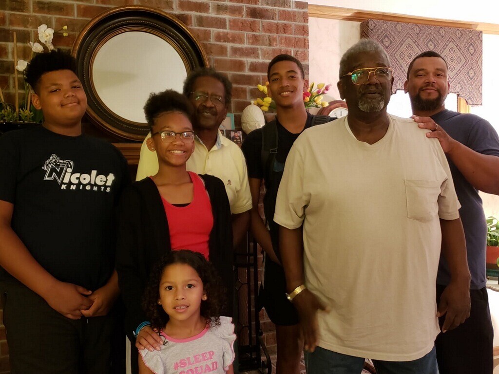 A group of seven people—three adults, three youths, and a small child—posing together in a living room with a brick fireplace and circular mirror behind them.