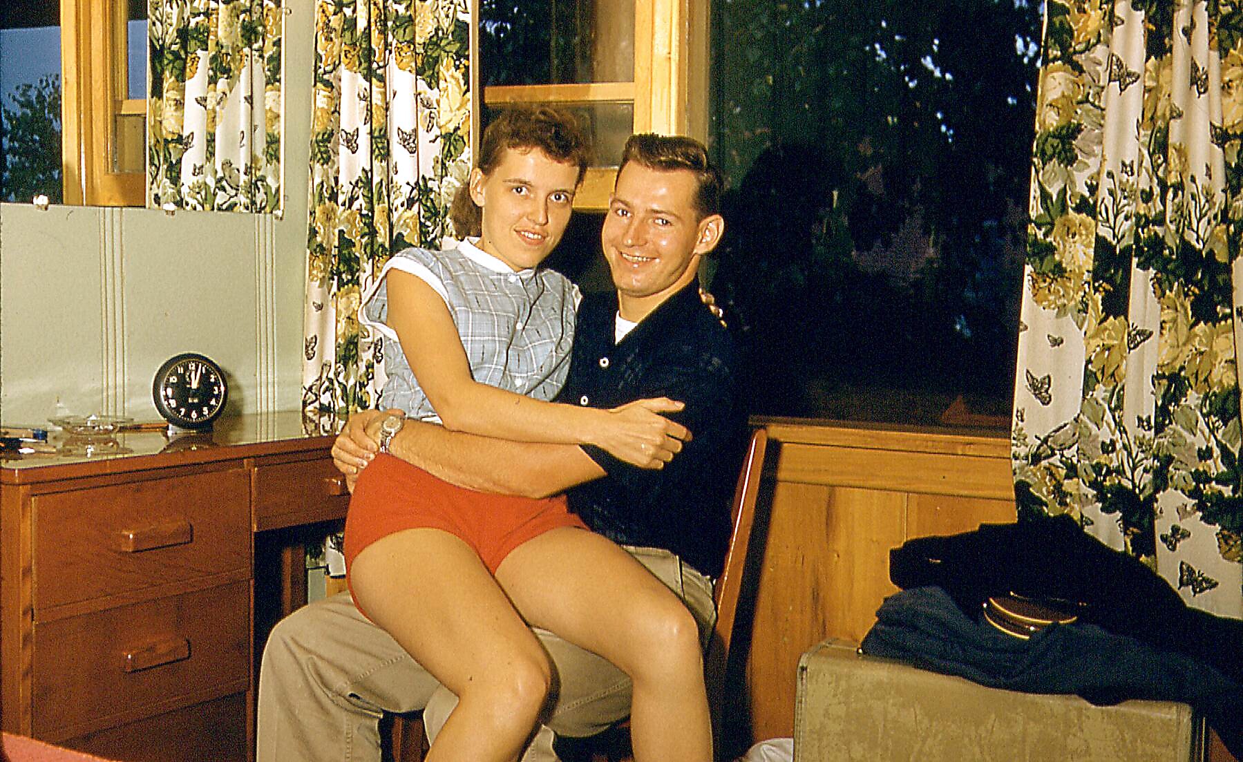 Couple posing together indoors; woman in blue plaid top and red shorts sits on man's lap by a wooden desk with a clock and butterfly-curtains in a vintage room.