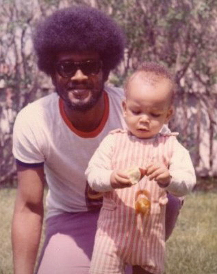 Man with an Afro and sunglasses kneels beside a toddler in striped overalls outdoors.