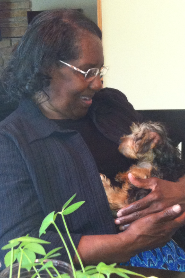 Woman wearing glasses smiling while holding a small fluffy dog in her arms, plant leaves in the foreground.