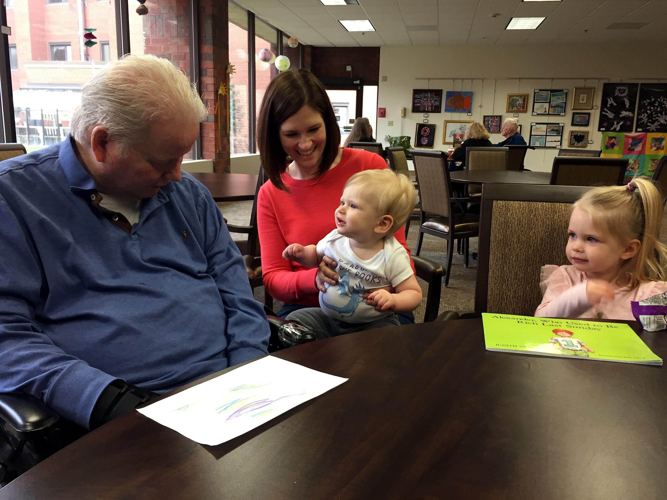 An older man in a blue shirt sits at a dark table while a smiling woman in a red top holds a baby; colorful wall art and windows visible in the background.