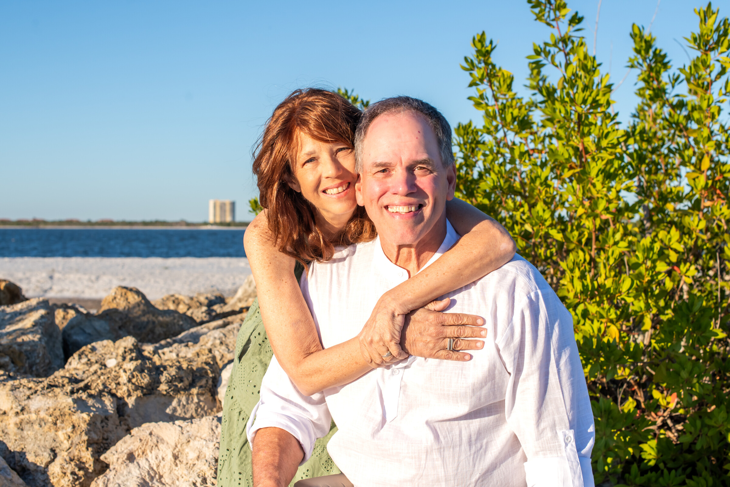 Smiling couple embracing on a rocky beach with blue water and green bushes in the background.