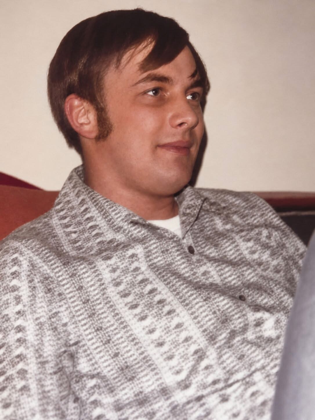 Man with brown hair and sideburns wearing a gray patterned sweater, seated indoors, looking to the right.