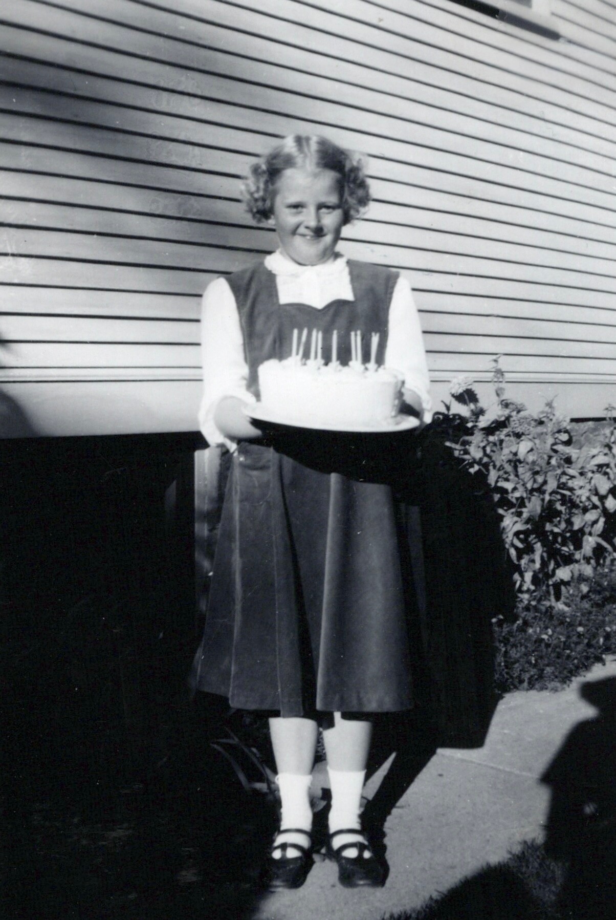 Girl in a vintage dress holding a round cake with candles in front of a wooden wall, celebrating a birthday.
