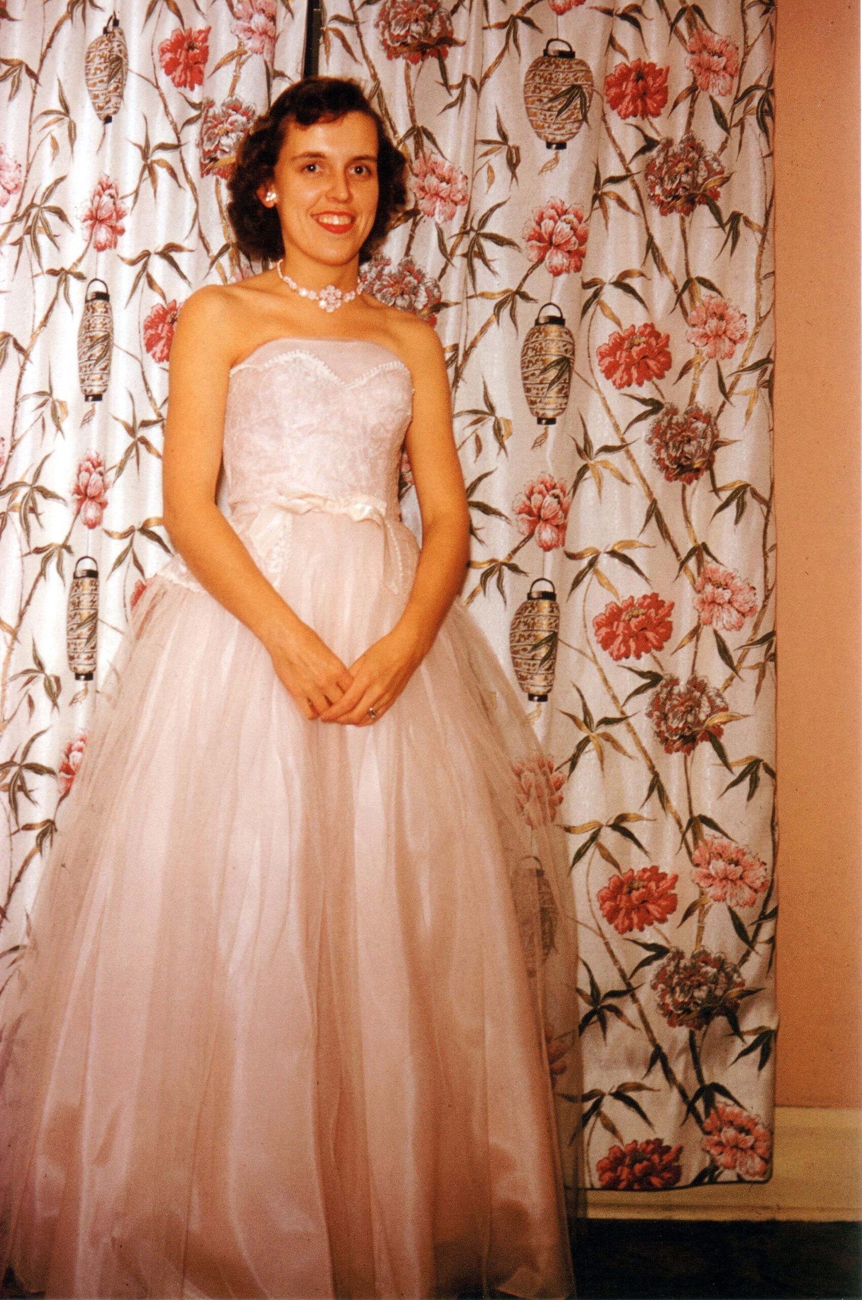 Smiling woman in a pale pink strapless wedding dress with a lace bodice and flower necklace, standing in front of a floral-patterned curtain.