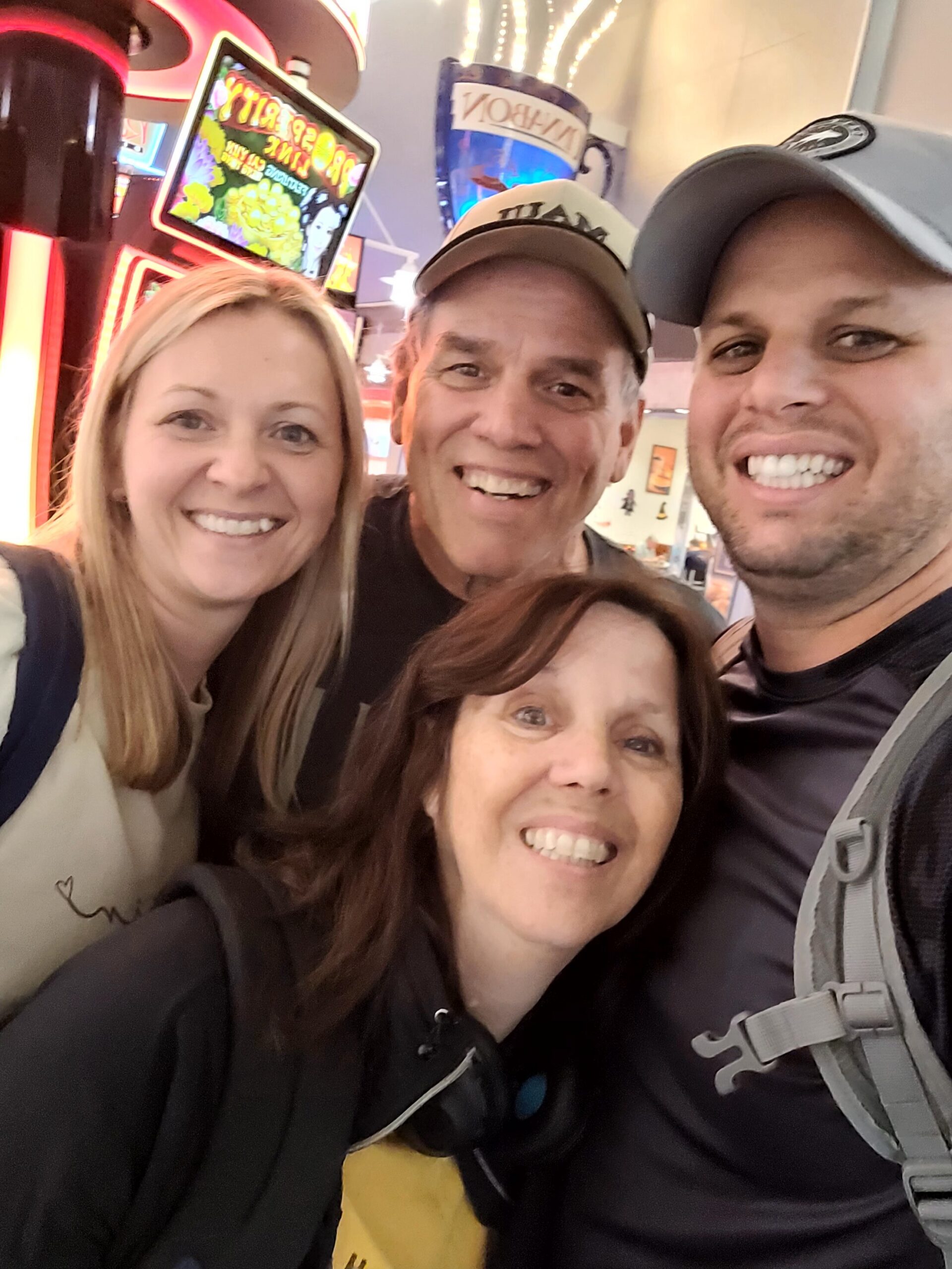 Four friends taking a close-up selfie indoors with colorful neon signs in the background, all smiling at the camera.