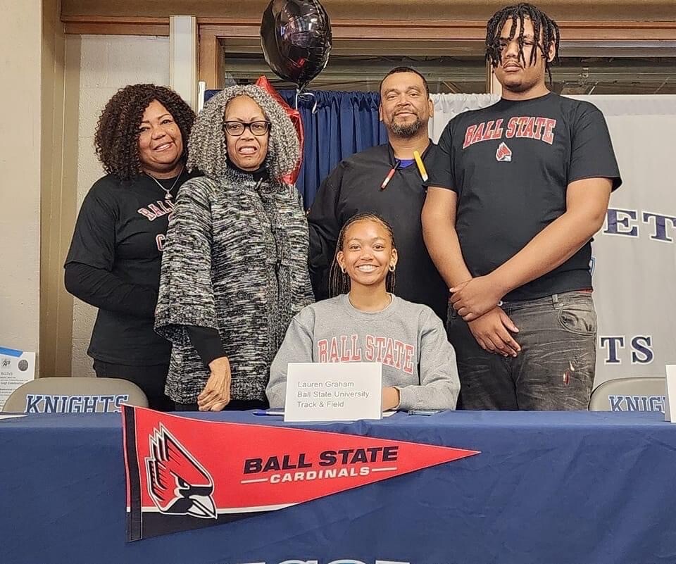 Group of five people posing for a signing day photo at Ball State, with a seated athlete wearing a Ball State shirt and a name card on the table.