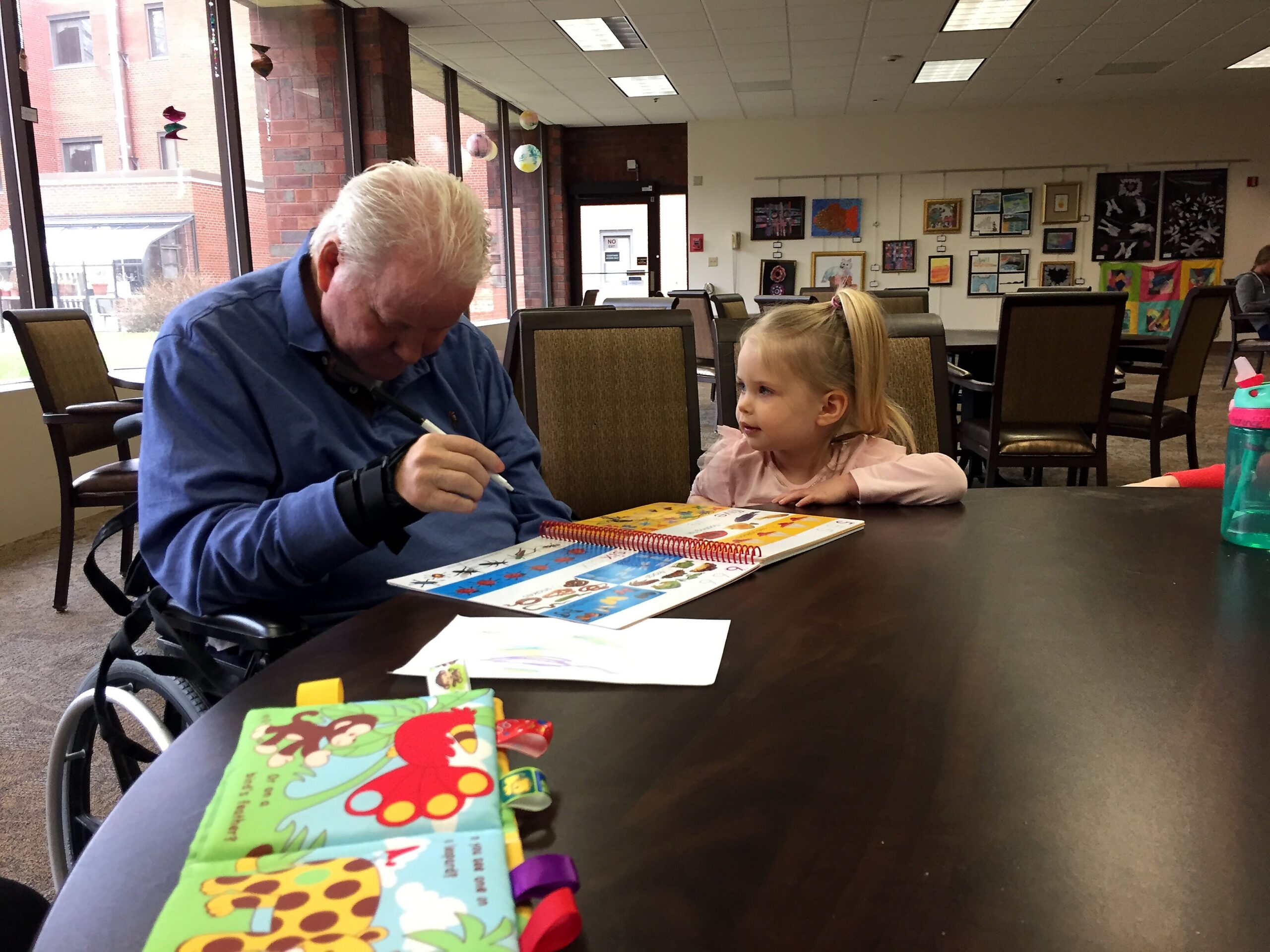 Older man in a blue shirt in a wheelchair sits at a table next to a young girl, looking at colorful activity books in a bright, art-filled room.