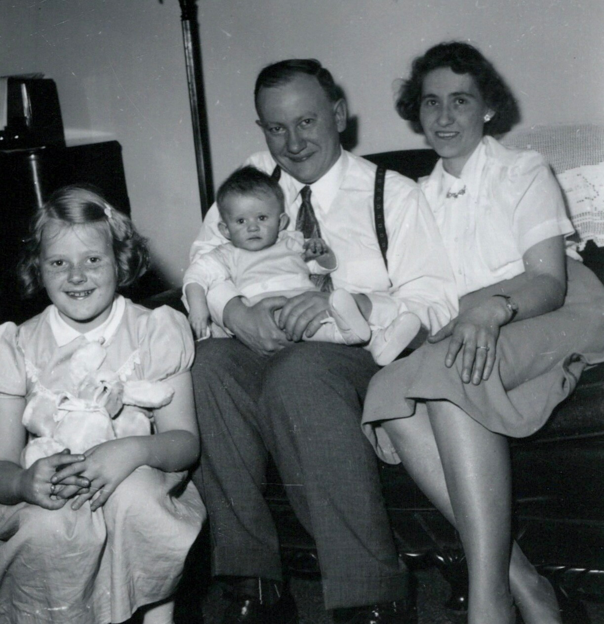 Family portrait on a couch: dad in a tie holding a baby, mom beside him, and an older sister smiling on the left in a white dress.