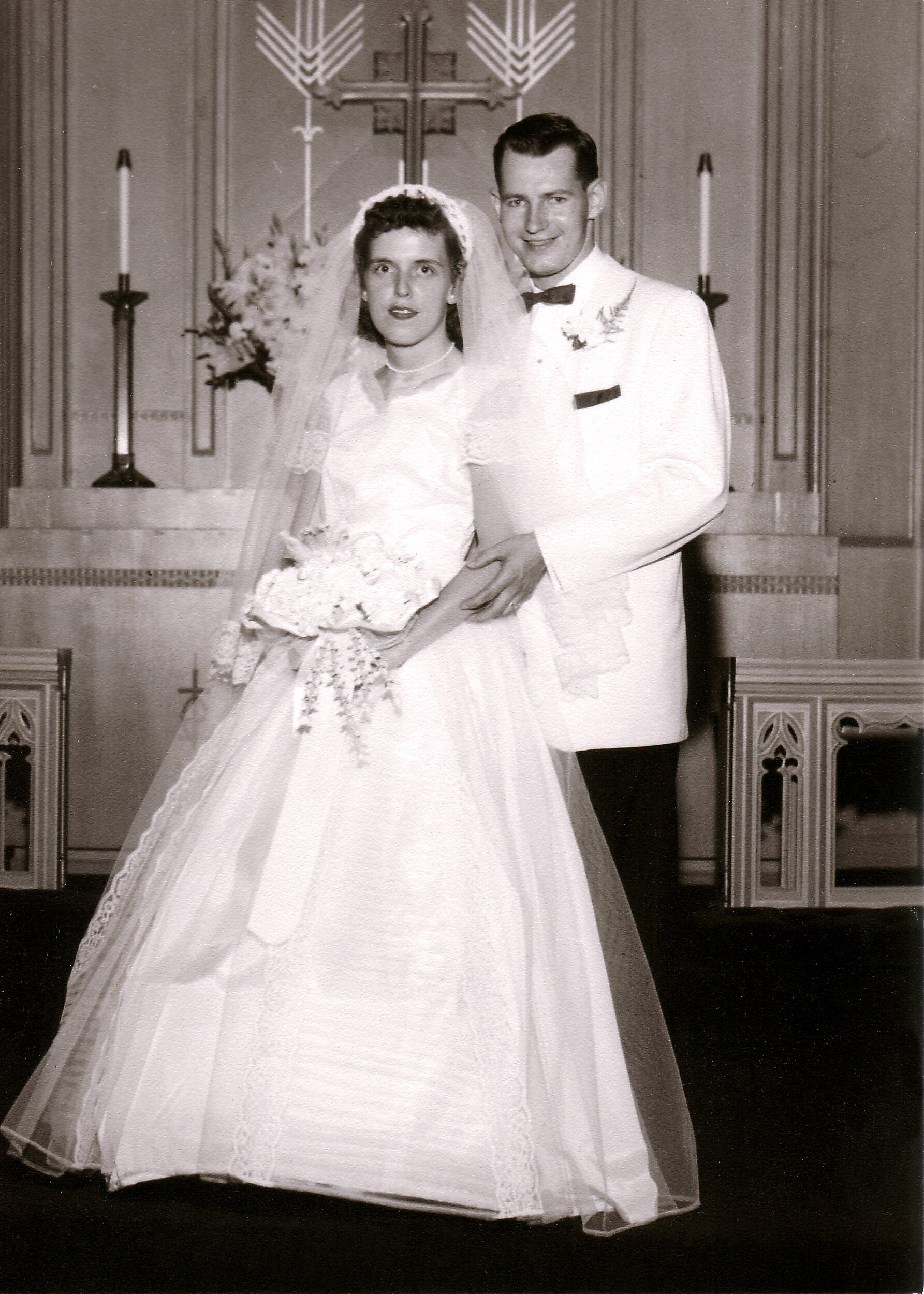 Bride in a wedding dress and veil stands with her groom in a white tuxedo at the church altar.