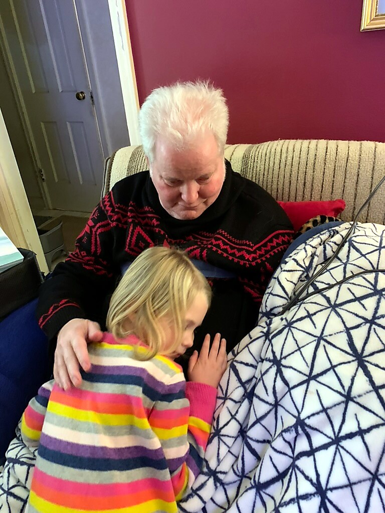 Tender moment: an elderly man with white hair sits on a sofa as a young child in a colorful striped sweater rests and leans against him under a cozy blanket.