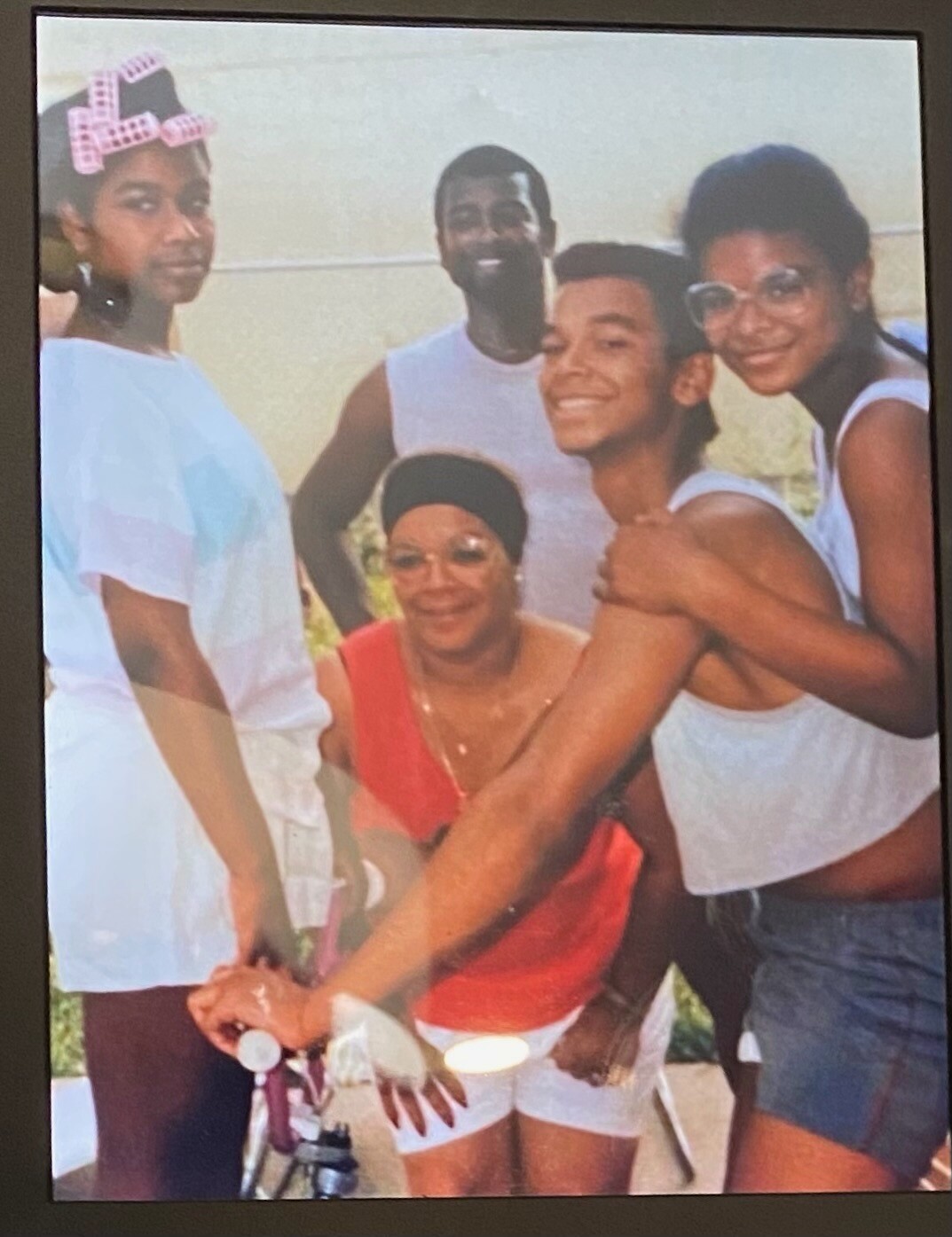 A group of five people posing outdoors for a photo: a woman in a red top kneels near a bicycle while four friends stand around, smiling and wearing casual white tops and light-colored clothing.