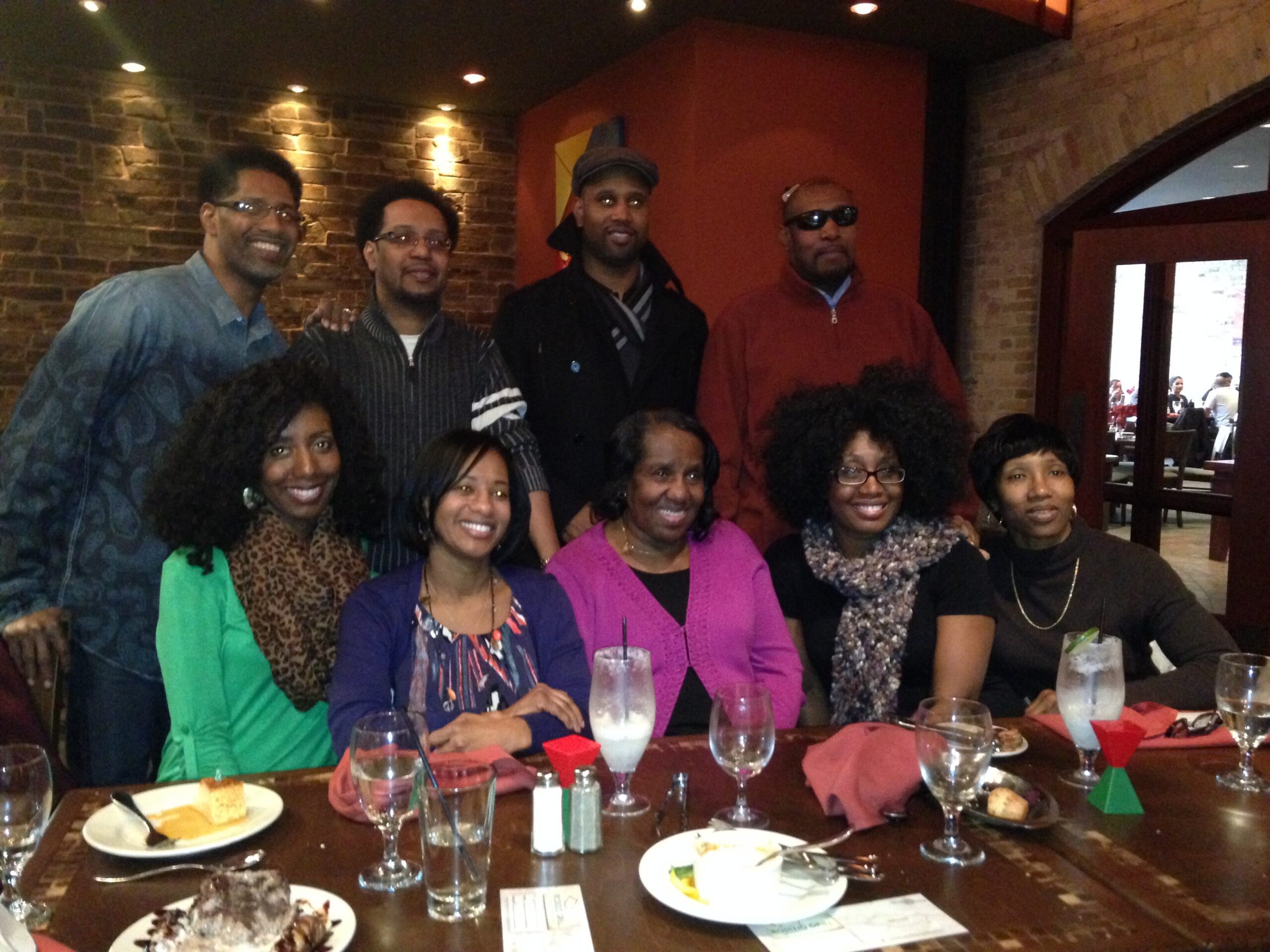 Group of diverse friends posing for a photo at a restaurant table, smiling with plates and drinks in front of them.