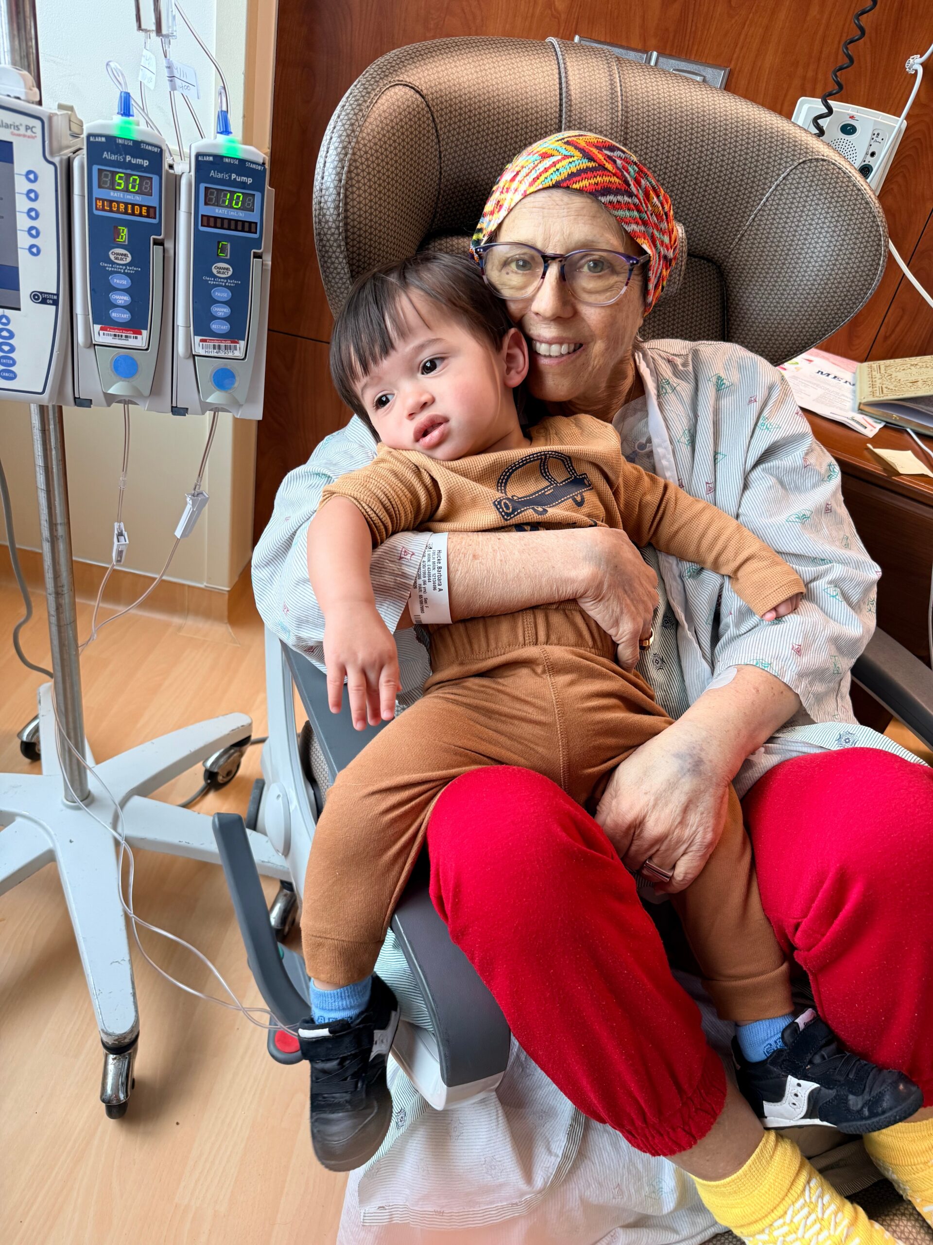 Older woman in a hospital chair smiling while holding a small child on her lap, with IV pumps visible nearby.