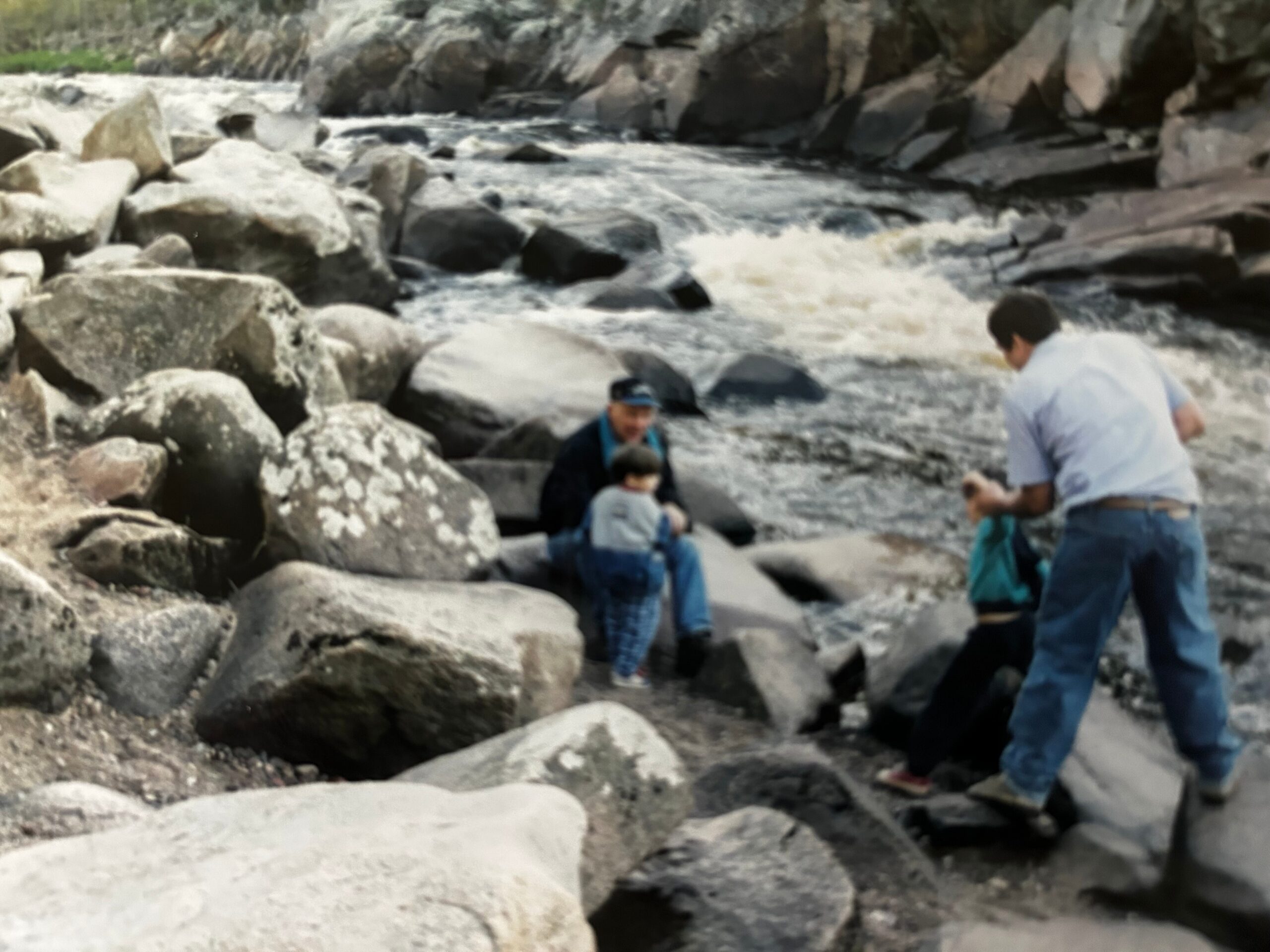 People exploring a rocky shoreline with large boulders and rough water in the background, possibly a family outing.