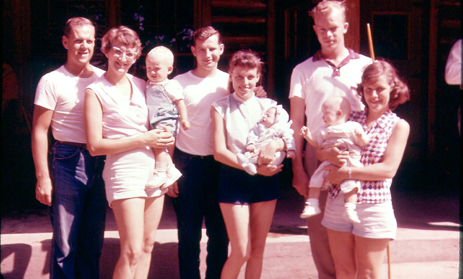 Group of adults and babies posing outdoors on steps, smiling in casual summer clothes.
