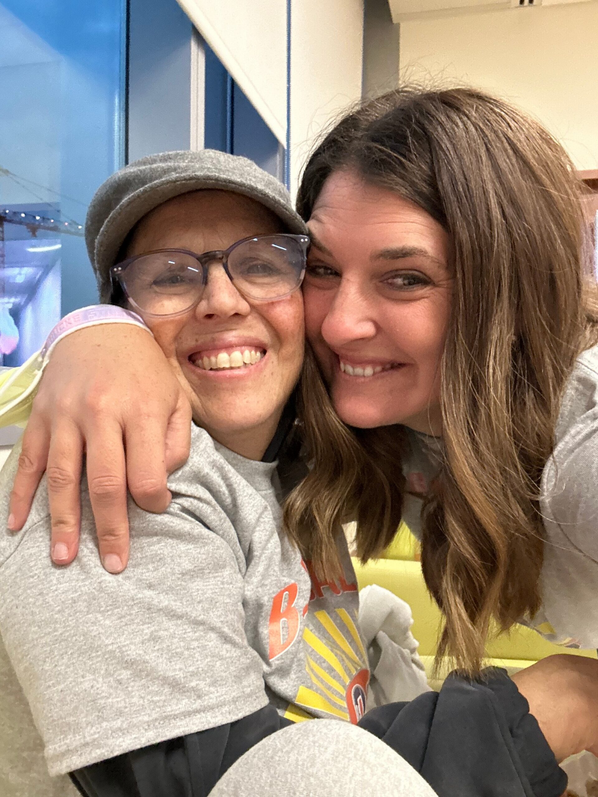 Two women smiling and hugging closely, one wearing a gray cap and glasses.