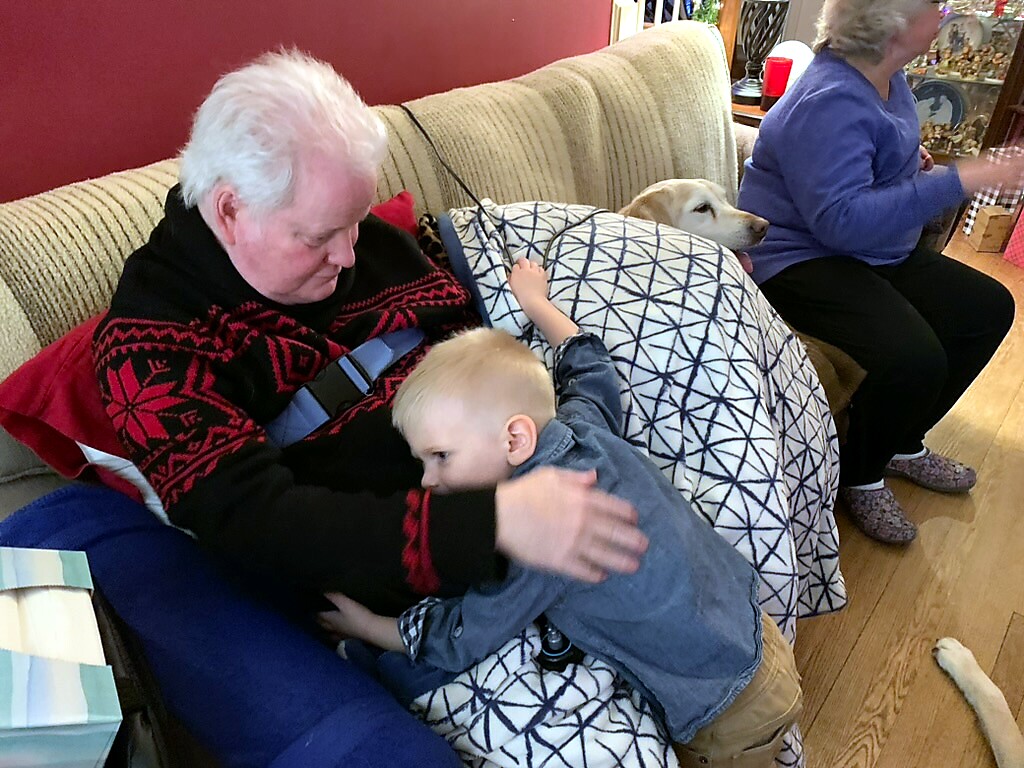 Grandfather on a couch being hugged by a young child, with a dog resting nearby in a cozy living room scene.