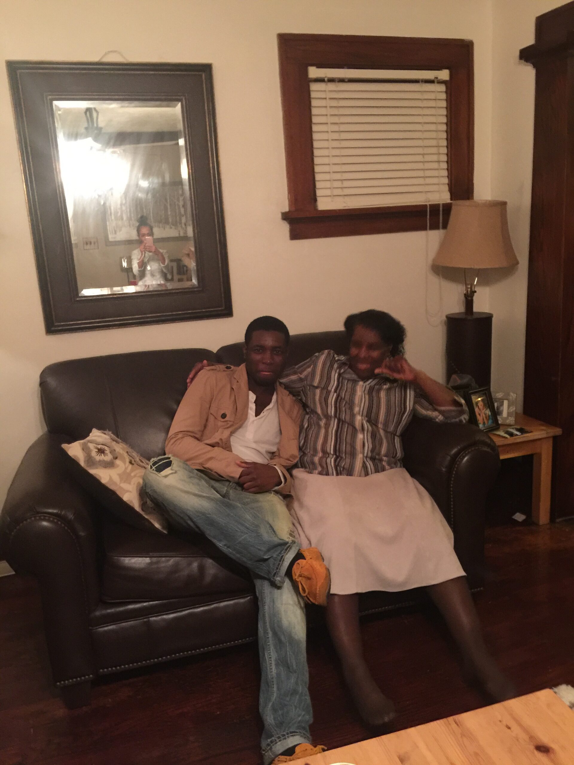 Two people sit close together on a dark leather sofa in a living room, smiling at the camera with one making a peace sign.