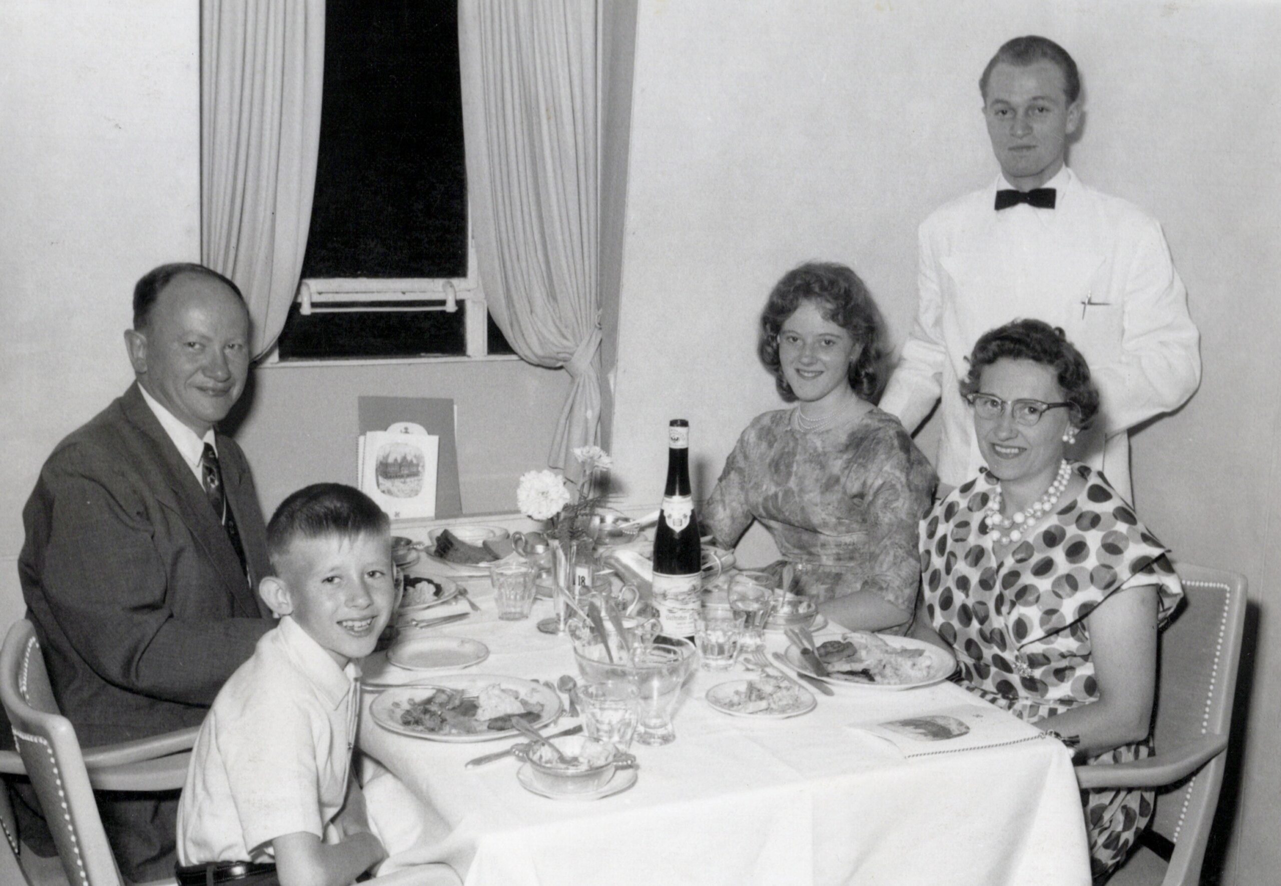 Black-and-white photo of a family at a formal dinner: two adults and two children seated, with a waiter standing behind them near the wall.