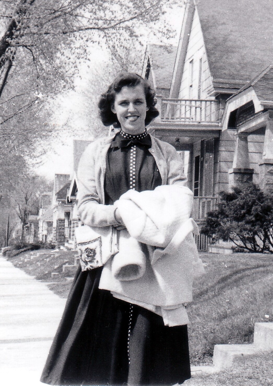 Woman in a vintage 1950s outfit standing on a suburban sidewalk, holding a light coat and a patterned handbag, smiling at the camera.