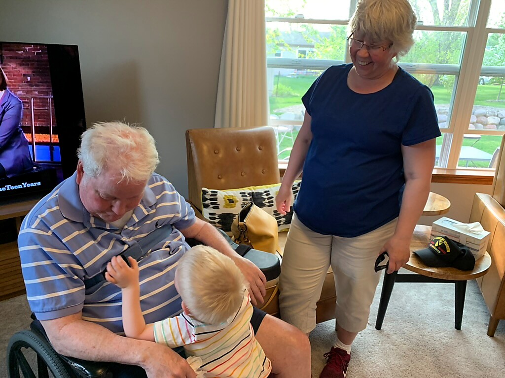 Older man in a wheelchair interacts with a toddler on his lap while a woman watches in a bright living room.